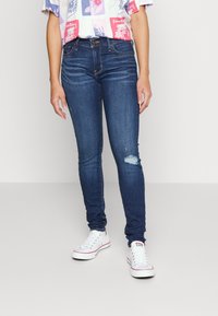 Jeans skinny in denim blu scuro con dettagli consumati sul ginocchio, abbinati a sneaker bianche. Il tessuto ha una texture liscia e leggermente elastica.