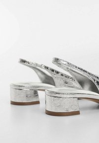 Srebrne, metaliczne sandały na słupku z teksturowanym wykończeniem, otwartym tyłem i delikatnym wygięciem. Posiadają solidną podeszwę z brązowym akcentem.