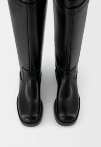 Par de botas de cuero negras altas con puntas cuadradas y acabado suave, de pie una al lado de la otra sobre un fondo blanco liso.