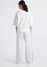 Woman with dark hair in a ponytail wearing a loose white half-sleeve top and light gray wide-leg pants standing against a plain white background.