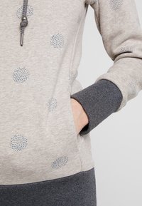 Handen i fickan på en beige huvtröja med blåa prickar och mörkgrå ribbade muddar och nederkant, vilket visar tygtexturen och snörknuten.