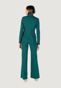 Una mujer está de espaldas al espectador, vistiendo un traje verde a medida y tacones beige, contra un fondo neutro.