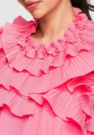 Vrouw draagt een felroze geplooide blouse met meerdere gelaagde ruches rond de halslijn en schouders.