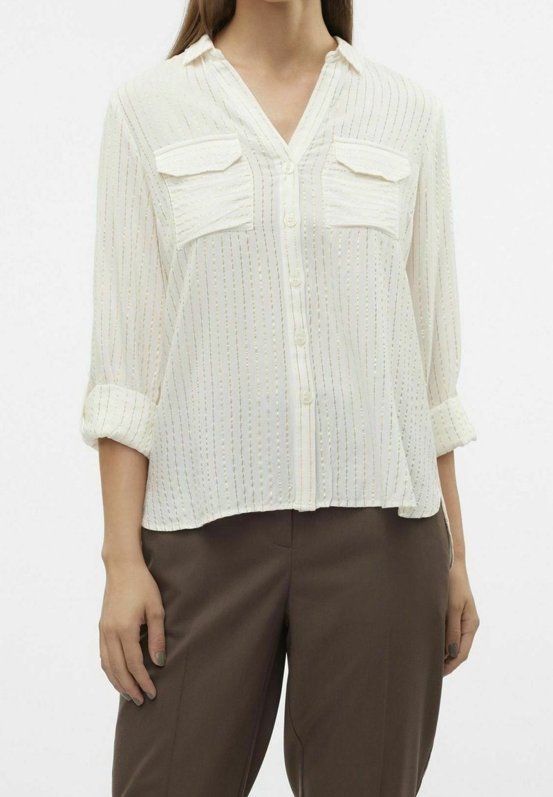 Femme portant un chemisier blanc transparent boutonné avec des rayures verticales dorées et deux poches poitrine à l'avant, associé à un pantalon marron.