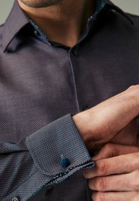 Sötétkék mintás ing kontrasztos gallérral és gombdíszítéssel. A közeli kép a texturált anyagot és a részletes mandzsettát mutatja kék gombbal.
