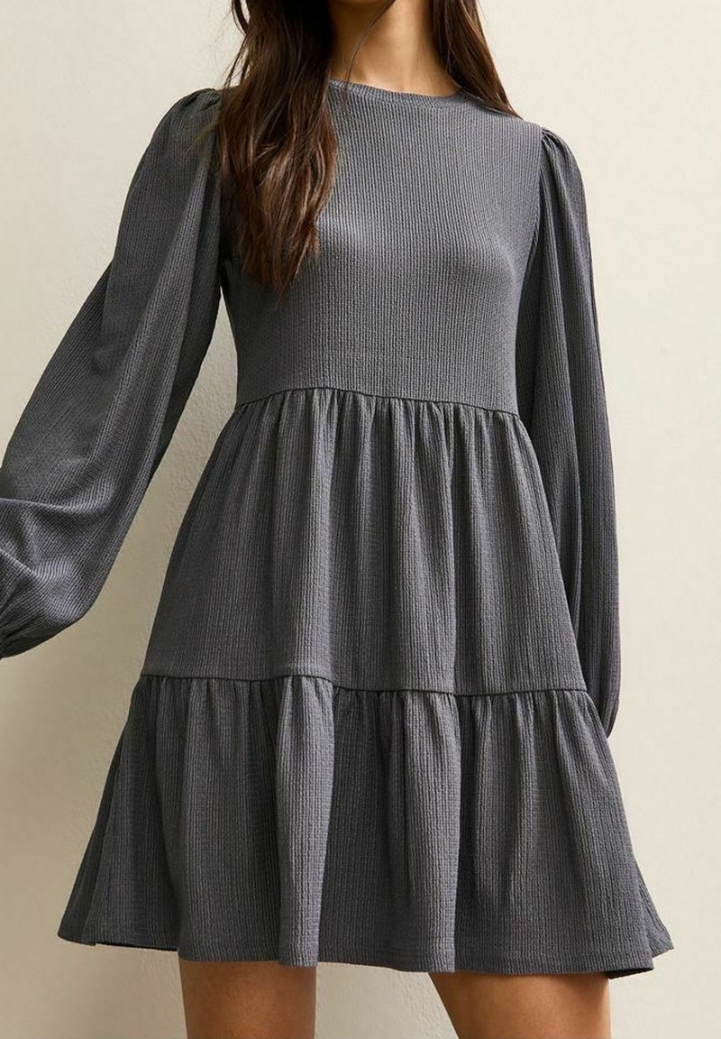 Femme portant une robe grise foncée à manches longues, à étages, avec un col rond et un tissu texturé.