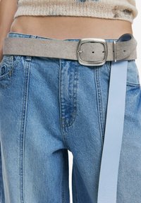 Gürtel aus grauem Wildleder mit einer quadratischen silbernen Schnalle, befestigt über blauen Jeans mit weitem Bein und sichtbaren Nähten.