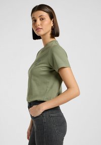 Zielona bawełniana koszulka z krótkim rękawem, okrągłym dekoltem, z subtelnym logo na klatce piersiowej. W połączeniu z ciemnoszarymi dżinsami.