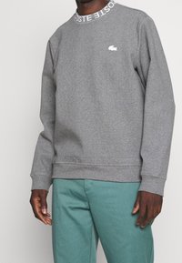 Man i en grå Lacoste-tröja med vitt logotyp och text vid halsringningen, i kombination med teal-gröna byxor.