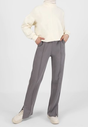 Person iført grå højttaljede bukser med frontsømme og sidelommer, cremefarvet sweatshirt og hvide sneakers, stående med hænderne i lommerne.