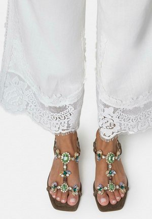 Pies con sandalias marrones de tiras en T decoradas con grandes gemas verdes y multicolores, combinadas con pantalones blancos con bordes de encaje.