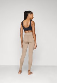 Svart sportsbh med brede stropper, kombinert med beige høye leggings som viser glatt tekstur og en tettsittende design. Ensfarget bakgrunn.