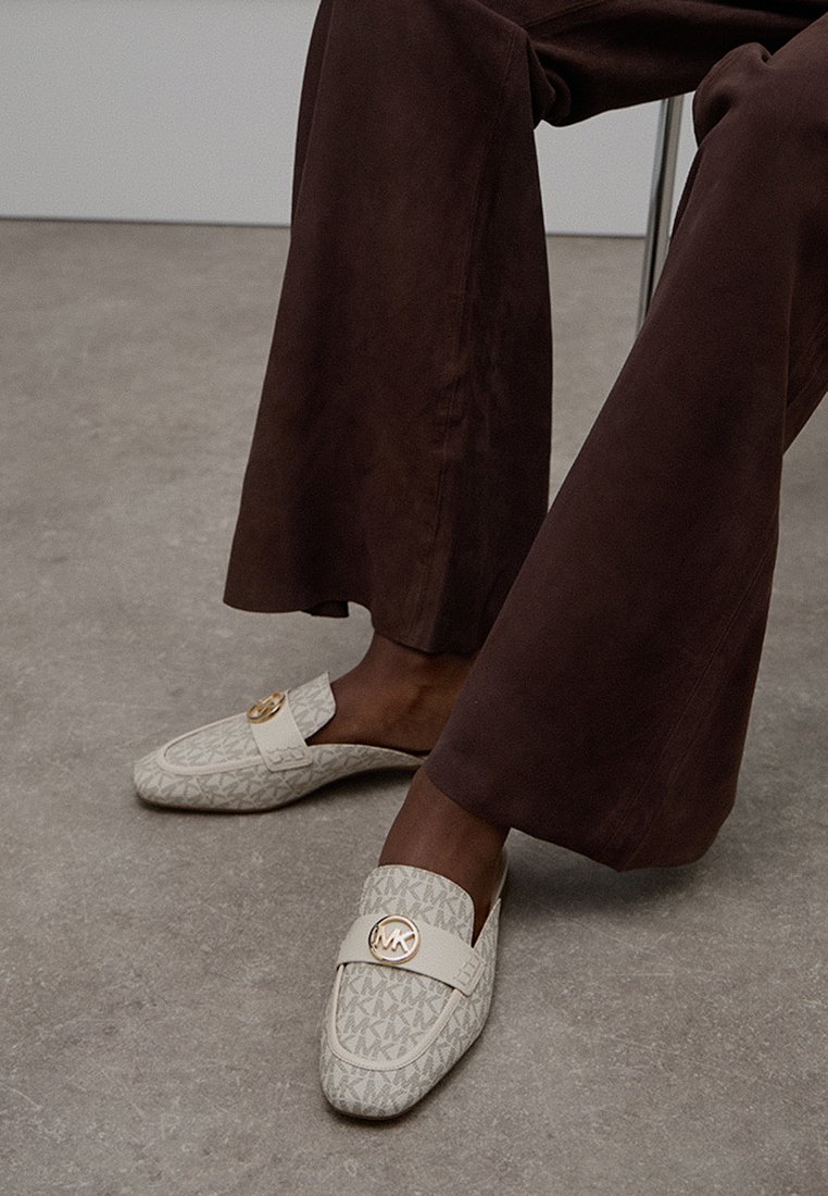 Beige instappers met een bovenwerk van een gemêleerd stof, voorzien van een gouden cirkelvormig logo accent. Gepaard met bruine wijde broekspijpen.
