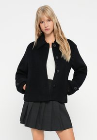 Žena u crnom vunenom sakoa s dugmadi preko bijele majice i tamno sivih plisiranih suknji, stoji s rukama u džepovima.