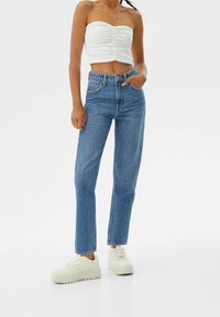 Jeans de mezclilla azul de cintura alta con corte recto, combinados con una blusa ajustada sin tirantes blanca y zapatillas de color claro.