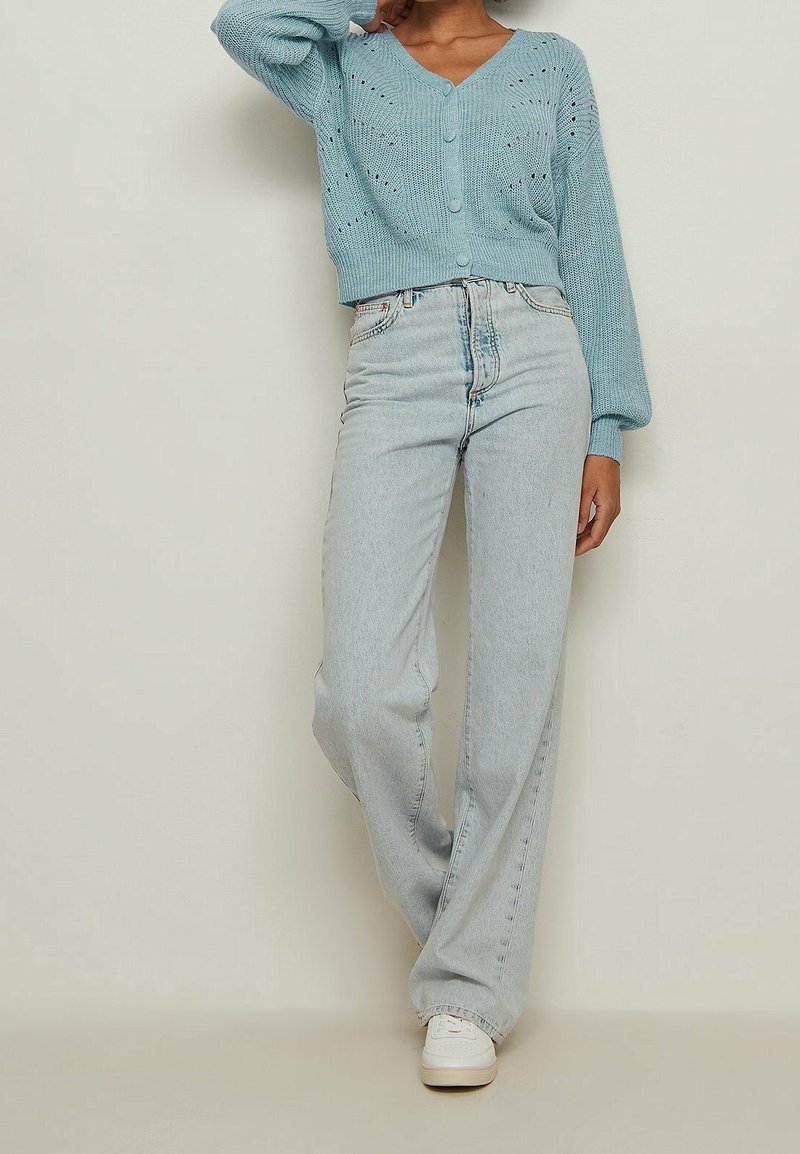 Person wearing light blue knit cardigan, high-waisted light wash jeans, and white sneakers, posing against plain beige background.