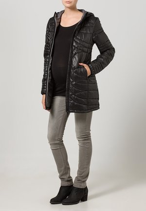 Chaqueta negra acolchada con capucha, que presenta un acabado brillante y un diseño acolchado. Llevada sobre una camiseta negra y jeans grises con botas hasta el tobillo.