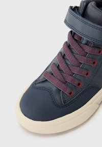 Høyhælte sneakers i marineblått med teksturert syntetisk materiale, røde og svarte flettede lisser, og en kremfarget gummisåle. Borrelåsstroppdetalj.
