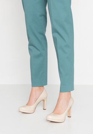 Underben iført blågrønne bukser sammen med beige højhælede pumps, der står på en glat hvid gulv.