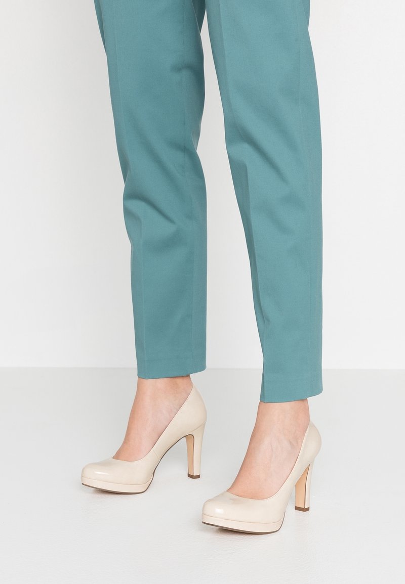 Jambes inférieures portant un pantalon turquoise associé à des escarpins beige à talons hauts, debout sur un sol blanc lisse.