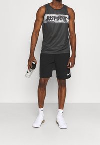 Canottiera sportiva grigia con grafica "JUST DO IT", abbinata a pantaloni neri e scarpe da ginnastica bianche. Bottiglia d'acqua tenuta nella mano sinistra.