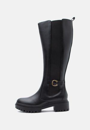 Schwarzer knöchelhoher Lederstiefel mit dicker Sohle, seitlichem Elastikeinsatz und dekorativer Messingschnalle auf weißem Hintergrund.
