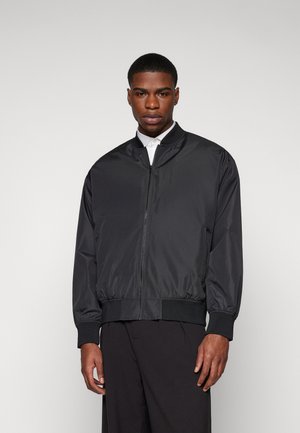 Giubbotto bomber nero con colletto alto, chiusura con zip e polsini elasticizzati. Realizzato in materiale leggero con una texture liscia.