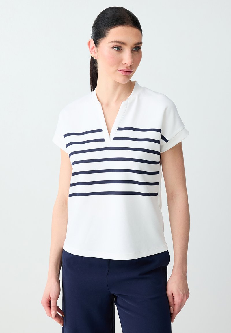 Femme aux cheveux foncés attachés en queue de cheval porte un haut blanc à manches courtes avec des rayures horizontales bleu marine et un pantalon bleu marine, debout devant un fond clair.