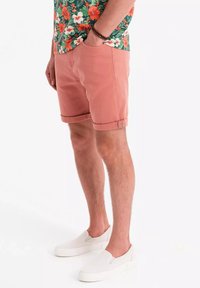Baumwollshorts in staubigem Rosa mit umgeschlagenen Säumen, kombiniert mit weißen Slippern. Das Modell trägt ein blumiges Hemd in Grün und Orange.