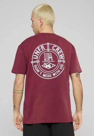 Mann mit kurzen blonden Haaren und Ohrringen, trägt ein kastanienbraunes T-Shirt mit weißem Text "UNFR CREW" und "DON'T MESS WITH US" mittig auf dem Rücken.