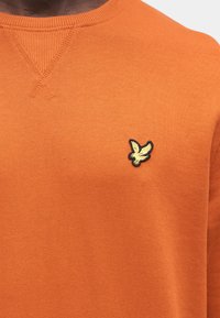 Orange sweatshirt featuring ribbed neckline, soft texture, and a black and yellow embroidered bird logo on the chest.