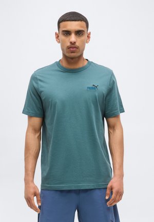 Jeune homme de face portant un t-shirt Puma bleu sarcelle et un short bleu sur fond gris clair uni.