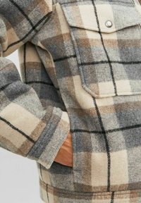 Rutet uldjakke med beige, brune og grå nuancer. Bemærkelsesværdige detaljer inkluderer en brystlomme og trykknap, samt en blød tekstur.