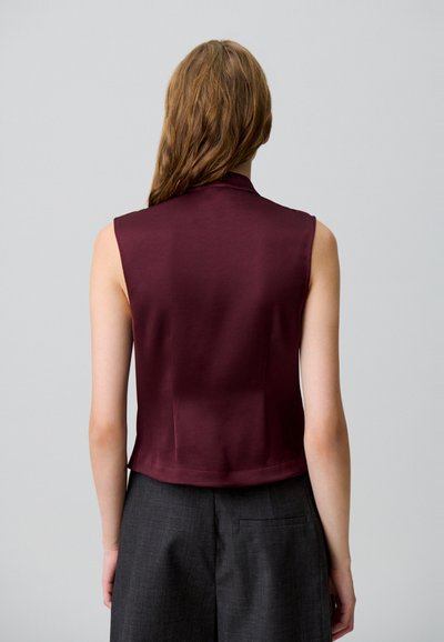 Top bordeaux senza maniche con colletto alto e texture liscia, caratterizzato da un design cropped, abbinato a pantaloni grigi scuri a vita alta.