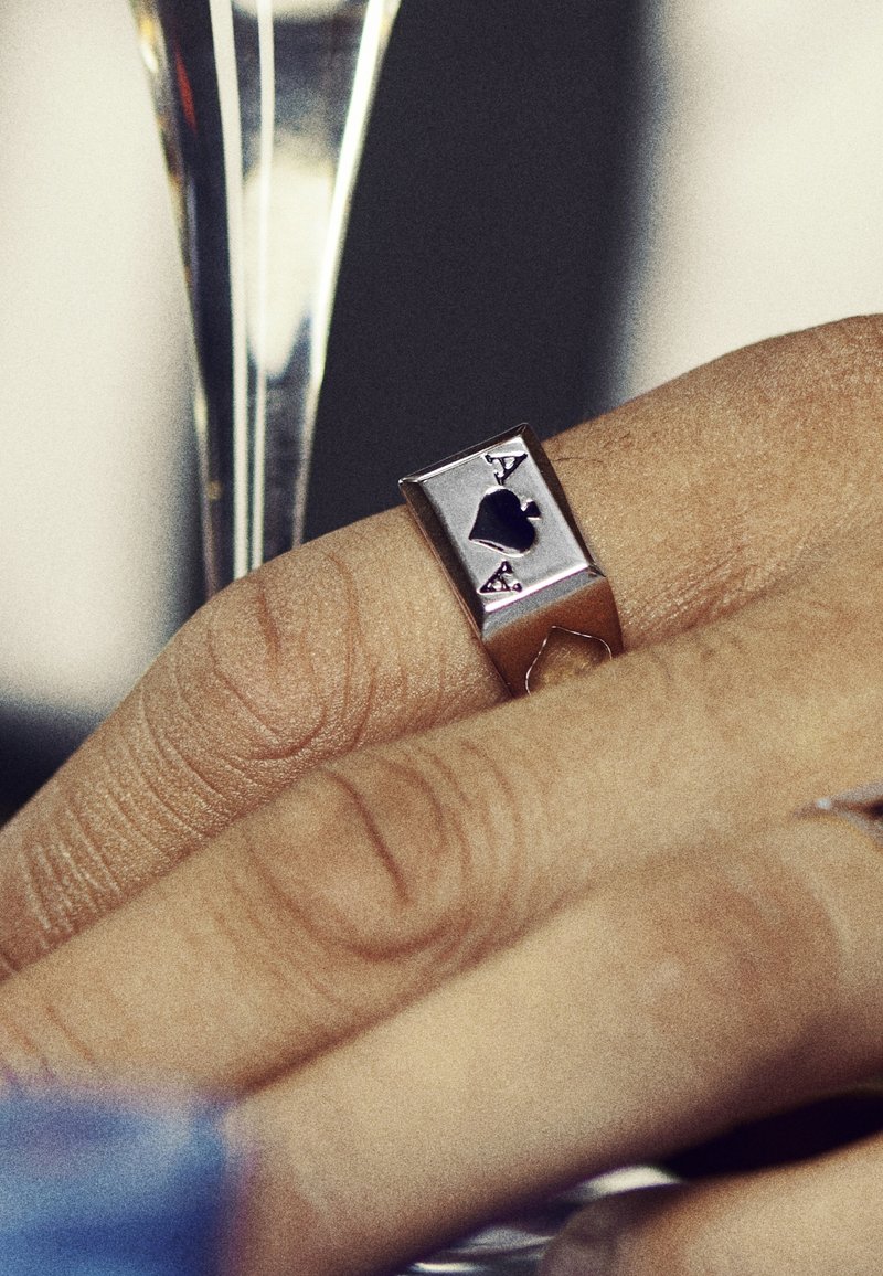 Silverring med rektangulärt mönster som har en präglad svart spader-symbol och "A", med en slät yta. Hand som håller ett glas.