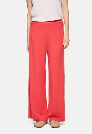 Mujer vistiendo pantalones anchos color coral con botones de madera a los lados y zapatos beige sin cordones, posando contra un fondo liso.