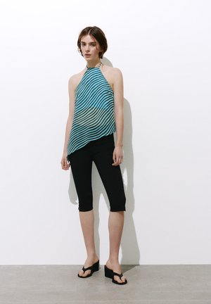 Donna con top halter a righe blu e nere, leggings neri al ginocchio e sandali con zeppa neri, in piedi contro un muro bianco.