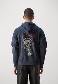 Felpa blu navy con grafica di un teschio accompagnato da rose e testo su banner sul retro. Realizzata in tessuto morbido, con un design rilassato e polsini a costine.