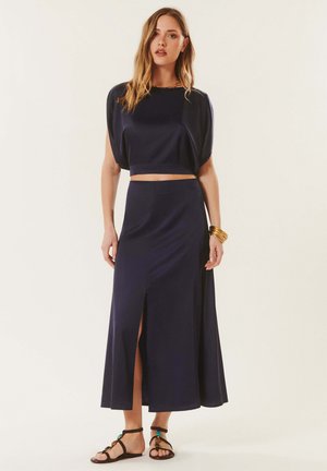 Femme portant un crop top bleu marine et une jupe taille haute assortie avec une fente devant, des bracelets dorés et des sandales plates noires avec des perles colorées.