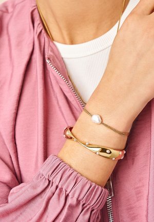 Handgelenk mit zwei Armbändern: eine goldene Kette mit einer einzelnen weißen Perle und ein breiteres goldenes Band mit kleinen Perlen, getragen über einer rosa Jackenmanschette.