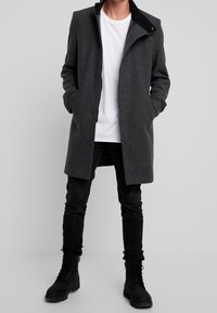 Cappotto di lana grigio con colletto alto, chiusure a scatto frontali e tasche laterali, indossato su una camicia bianca e jeans neri, abbinato a stivali neri.