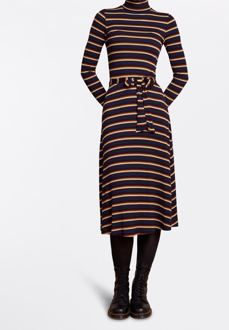 Robe côtelée bleu marine avec des rayures horizontales jaunes et rouges, manches longues, col roulé, ceinture à la taille, associée à des collants noirs et des bottines à lacets.