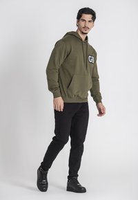 Felpa verde oliva con tasca frontale e logo "GK" bianco, abbinata a jeans neri e sneakers nere. Fit casual, texture liscia.