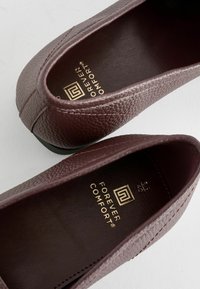 Burgundské slip-on topánky s textúrovaným koženým exteriérom. Interiér obsahuje zlaté logo s nápisom "FOREVER COMFORT" na tmavej koži.