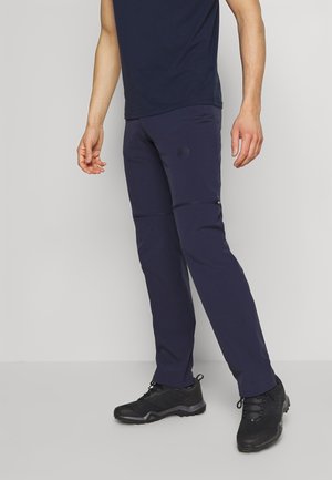 Mężczyzna ubrany w granatowe spodnie trekkingowe z odpinanymi nogawkami i czarne sportowe buty stojący na jasnym, jednolitym tle.