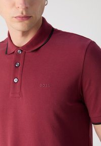Bordová polo košeľa s jednostrannou textúrou, čiernymi akcentmi na golieri, tlačidlami na zapínanie a vyrazeným logom "BOSS" na hrudi.