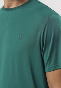 Groen T-shirt met ronde hals, gemaakt van gladde stof. Heeft een subtiel logo in een donkerdere tint, korte mouwen en een aansluitend ontwerp.