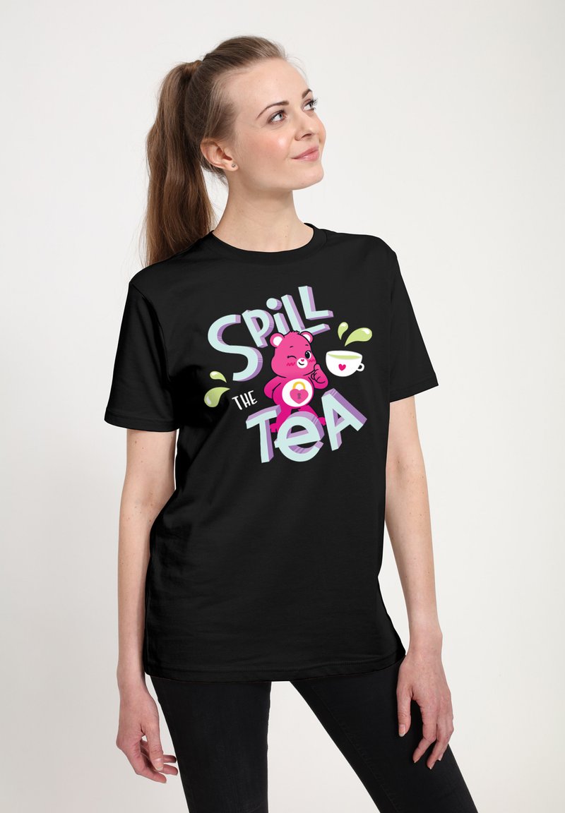 Camiseta negra con un diseño gráfico que presenta un oso rosa, la frase "Spill the Tea" y una taza de té con salpicaduras. Manga corta, ajuste casual.