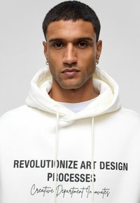 Witte hoodie met zwarte tekst "REVOLUTIONIZE ART DESIGN PROCESSES" en "Creative Department Innovates," met een trekkoordkap.