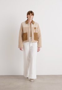 Veste en shearling beige avec poches en daim marron, détail en anneau doré et col. Associée à un pantalon large blanc sur un fond neutre.